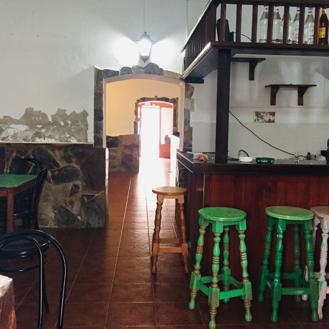 Interior de un restaurante con paredes de piedra, mesas con sillas y un bar de madera verde, luz brillante en la entrada y algunas botellas en la estantería.