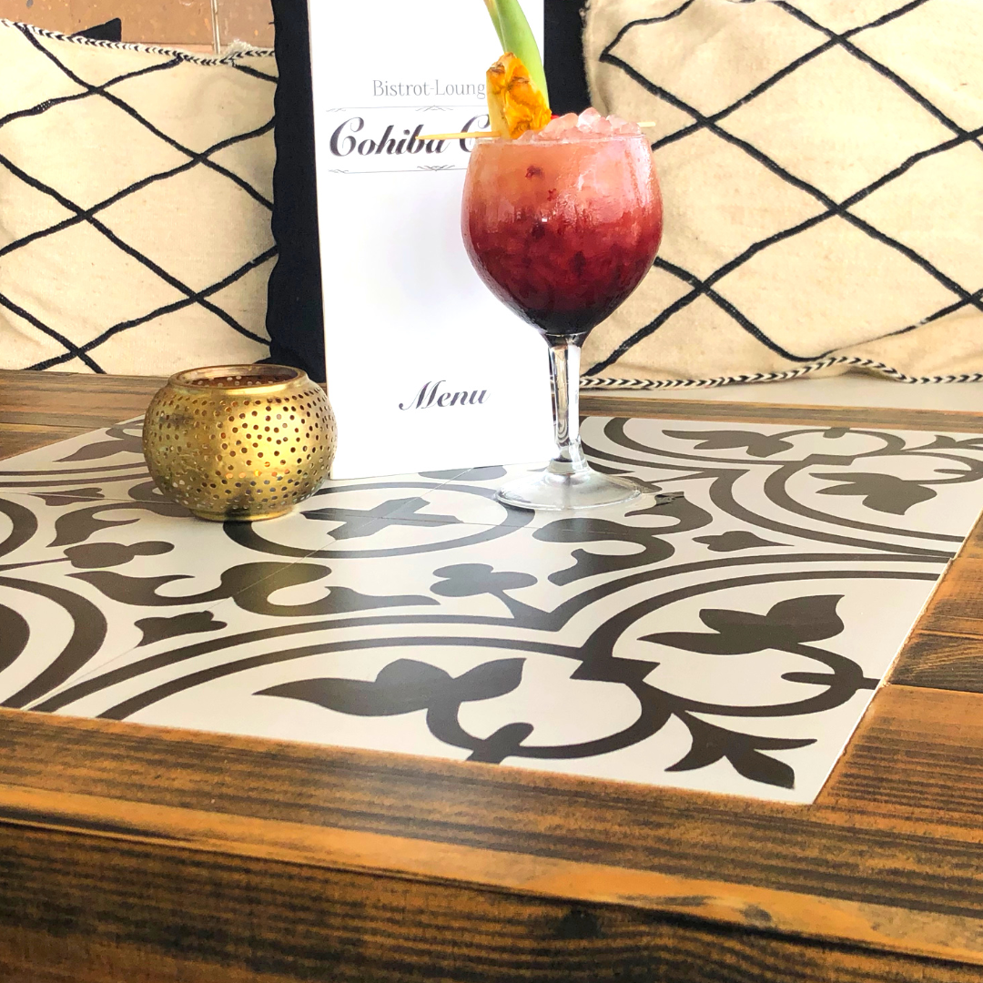 Vaso con cóctel de color rojo y rosa, decorado con piña y una hoja verde, en una mesa de madera con mantel decorativo negro y blanco, en un restaurante o bistró.