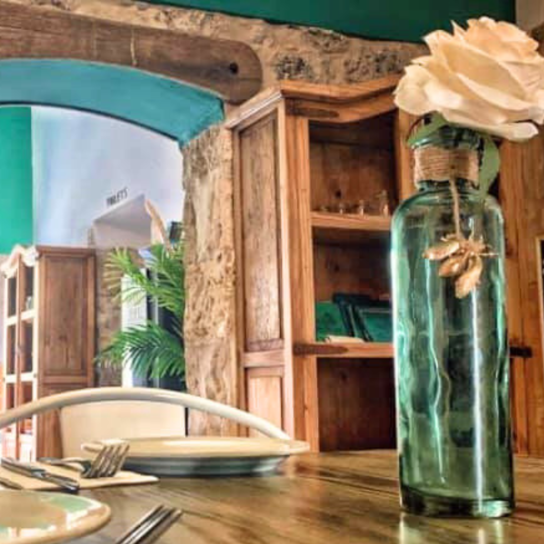 Interior de un restaurante con una mesa de madera, plato, tenedores, y un florero con flores en primer plano, y estanterías de madera y plantas decorativas en el fondo.