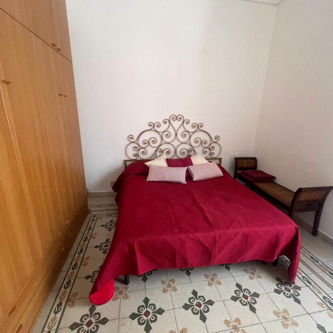 Cama con cabecera de estilo de hierro forjado y ropa de cama roja, almohadas a rayas y de color sólido, habitación con armario de madera a la izquierda y banca con cojines a la derecha, piso de azulejos decorados.