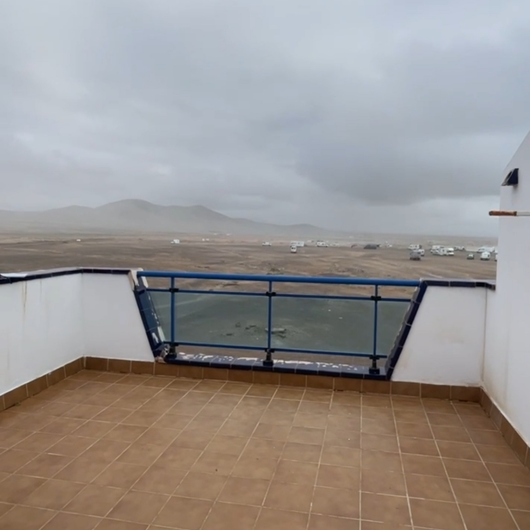 Vista desde una terraza con suelo de baldosas, mostrando un paisaje árido con colinas y varias caravanas o casas móviles en la distancia, bajo un cielo nuboso.