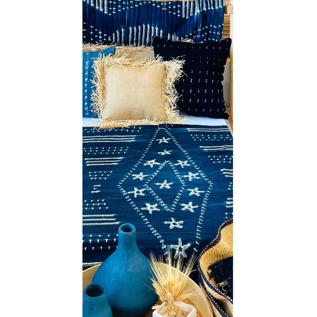 Ropa de cama azul con bordados blancos, con cojines negros y beige con flecos. Decoración con vasijas y plantas secas en un entorno interior.