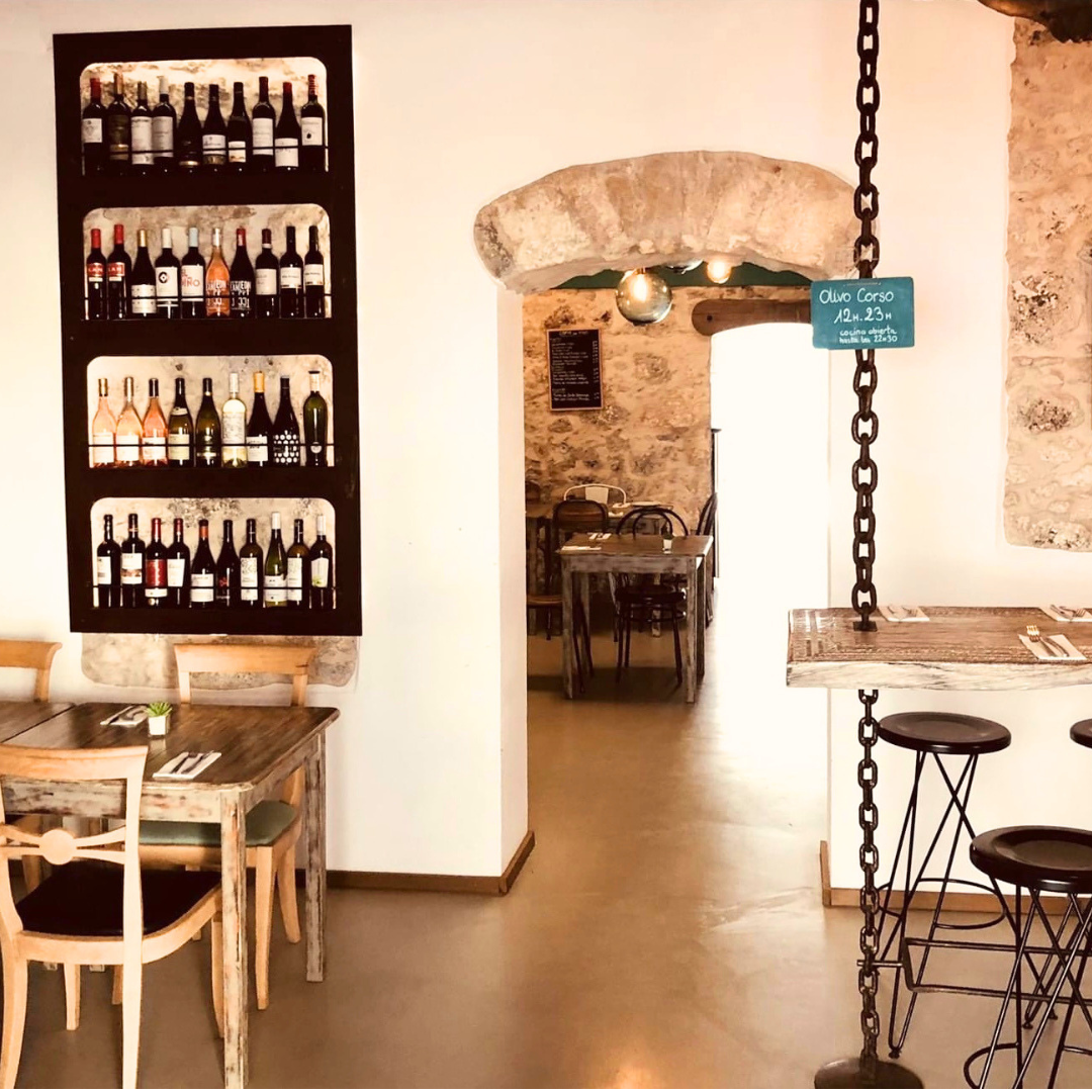 Interior de un restaurante con paredes de piedra, una estantería con botellas de vino, mesas de madera y sillas, y banderines colgantes de cadenas.