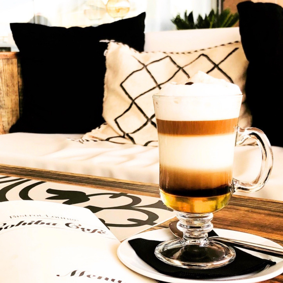 Vaso de café con leche y espuma en un café o restaurante, con fondo de cojines decorativos en un sofá, uno negro y uno blanco con diseño negro.