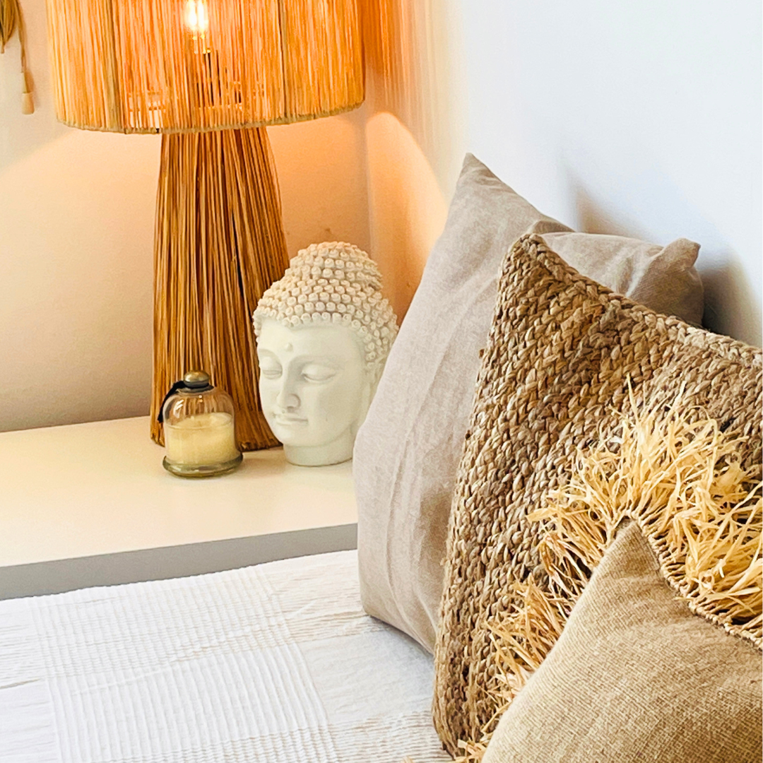 Alfombra blanca, cojines beige y marrón en la cama junto a una lámpara de madera, una cabeza de buda blanca y una vela en una mesita de noche.