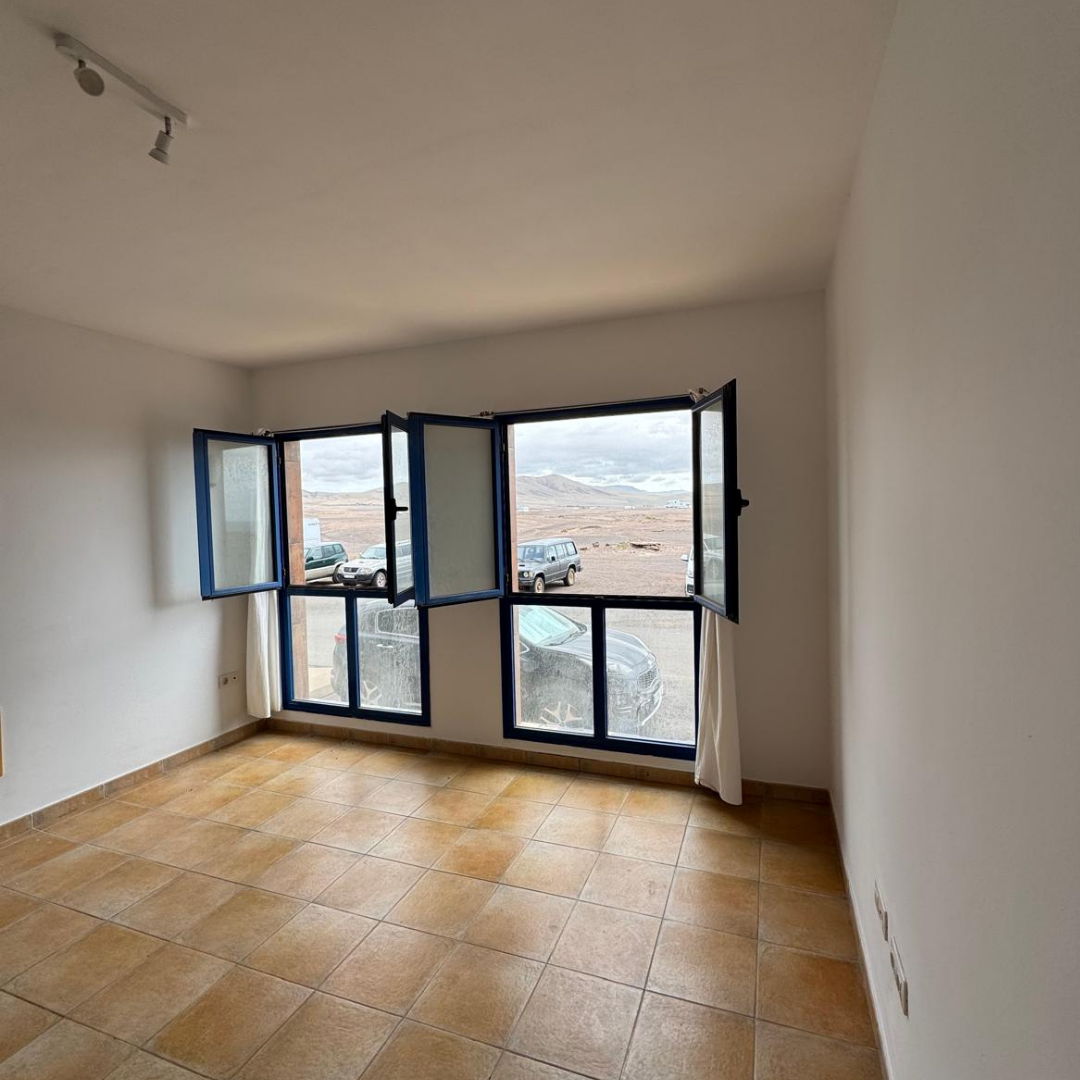Habitación vacía con ventanas abiertas y vista a un paisaje desértico, con autos estacionados afuera y piso de baldosas beige.