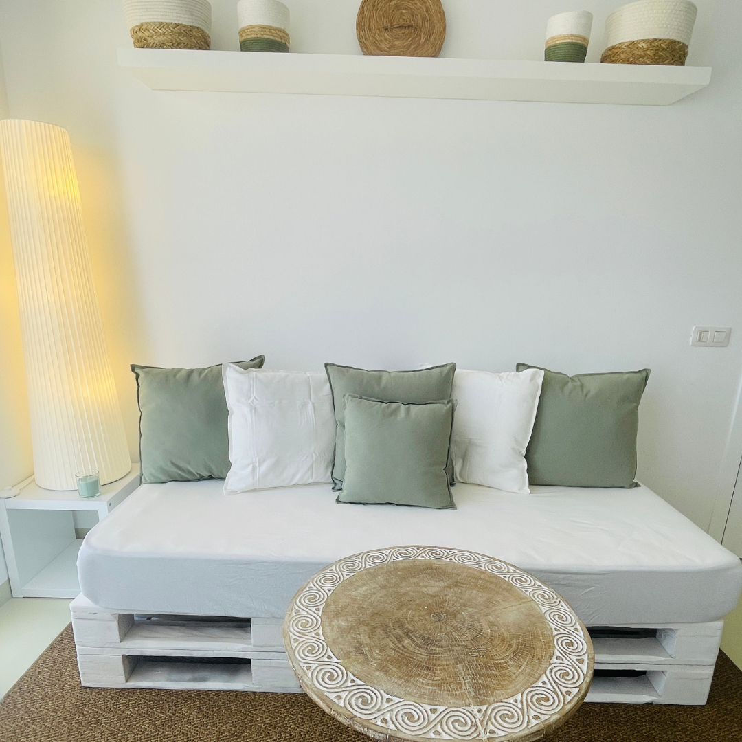 Sofá blanco con siete almohadas verdes y blancas, mesa redonda de madera con grabados, lámpara de piso amarilla, estantería con varias cestas decorativas, alfombra marrón.