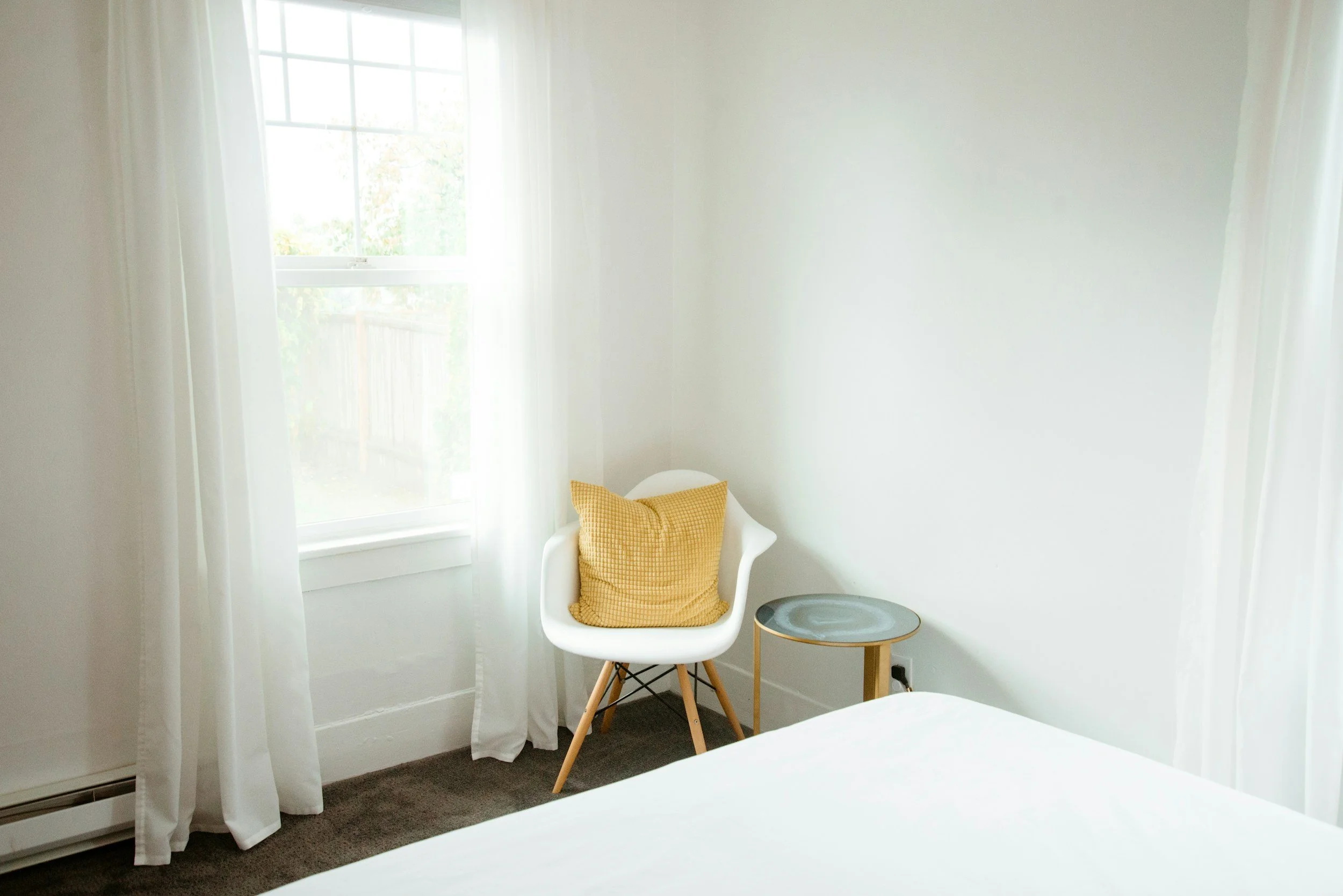 A corner of a bedroom with a white armchair that has a yellow checked cushion, a small round side table, a window with white curtains, and a bed with white bedding.