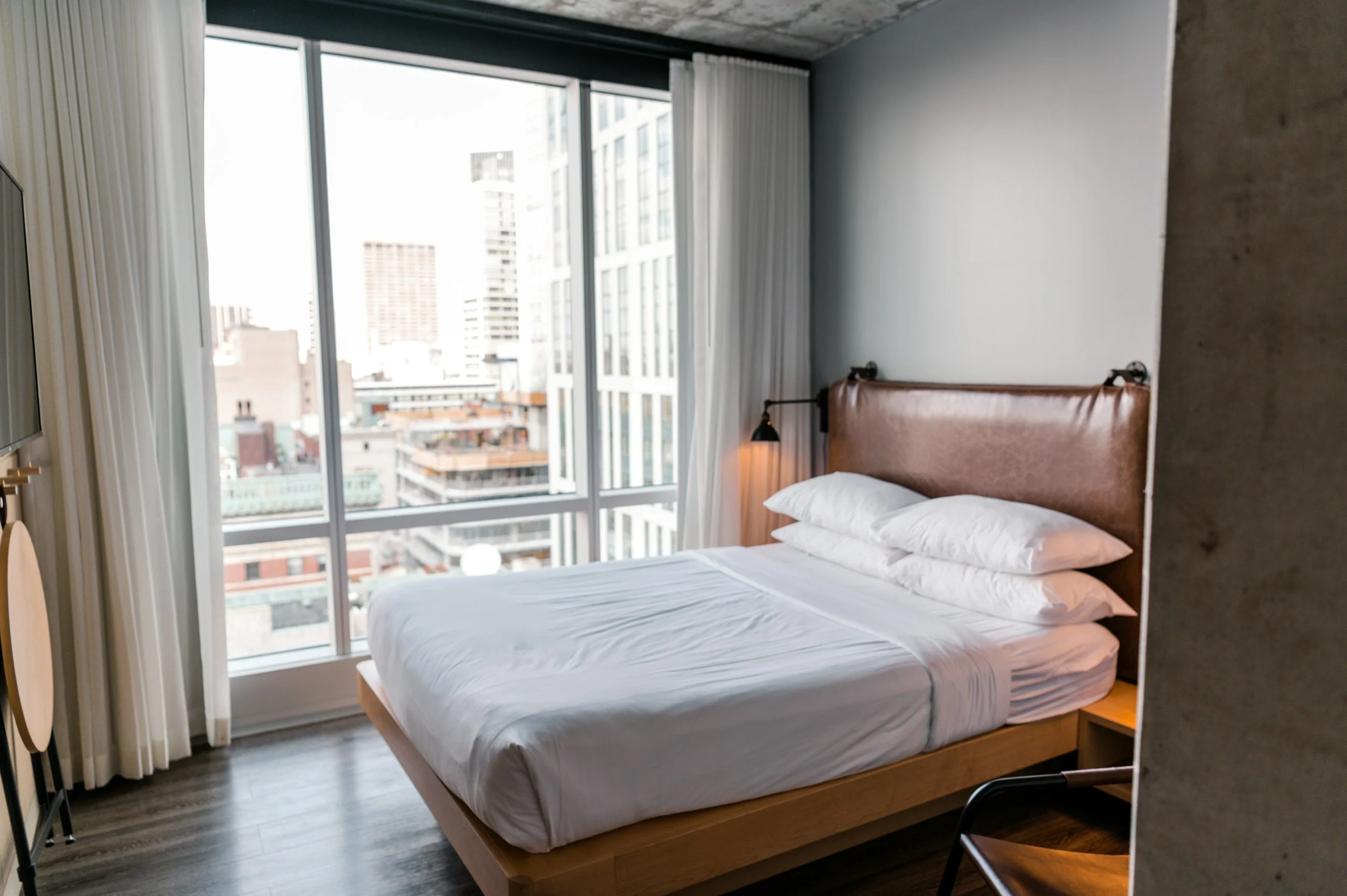 A modern hotel room with a large bed, white bedding, and a leather headboard. Floor-to-ceiling windows with a city view and white curtains. A bedside lamp and part of a chair are visible.