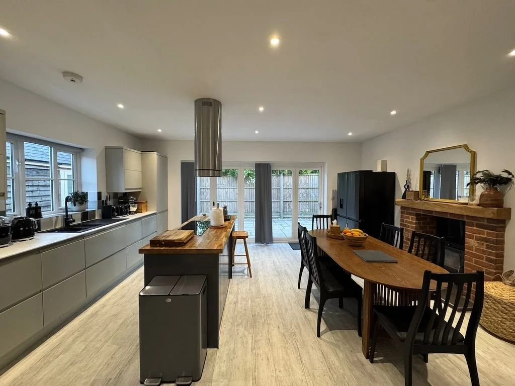 Modern kitchen and dining area with sliding glass doors leading to an outdoor patio, featuring a kitchen island, dining table with six chairs, brick fireplace with mirror, and various kitchen appliances.