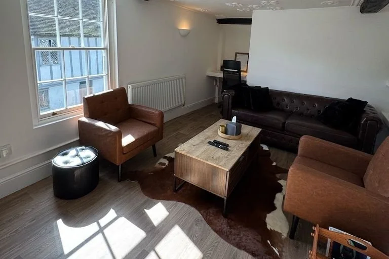 A cozy living room with a window letting in natural light, two brown armchairs, a black leather sofa, a wooden coffee table with remote controls and a tissue box, a cowhide rug, a small black ottoman, a white radiator, and a desk with a chair in the corner.
