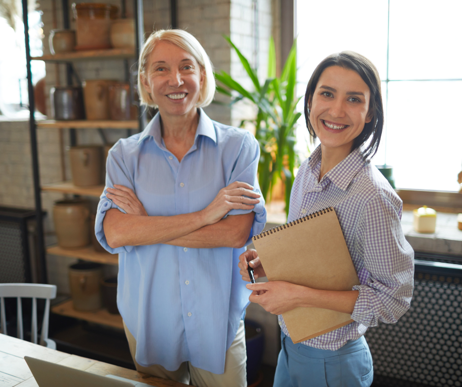 Two women smiling in an office environment, with the younger woman holding a notebook and pen and the older woman crossing her arms.