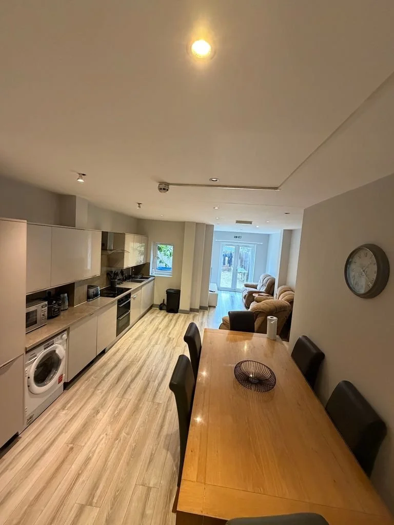 Interior view of an open-plan living space with a kitchen, dining area, and sitting area, featuring wooden flooring, modern appliances, and cozy chairs.