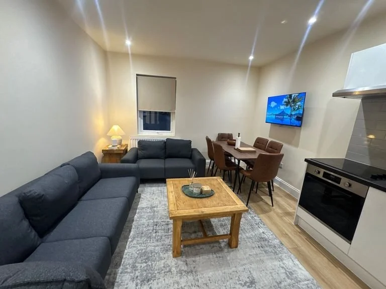 Living room with two dark gray sofas, a wooden coffee table, a dining table with six brown chairs, a wall-mounted TV showing a beach scene, a window with a blind, a small side table with a lamp, and a stove in the corner.