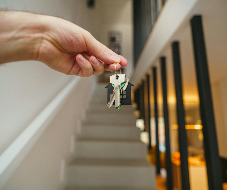 A person holding a keyring with a house-shaped keychain and house keys in front of a staircase and a railing inside a home.