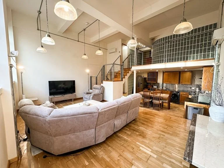 Open-concept living room and kitchen with hardwood floors, beige sectional sofa, TV, armchair, dining table with chairs, kitchen cabinets, and appliances, with a loft space above.