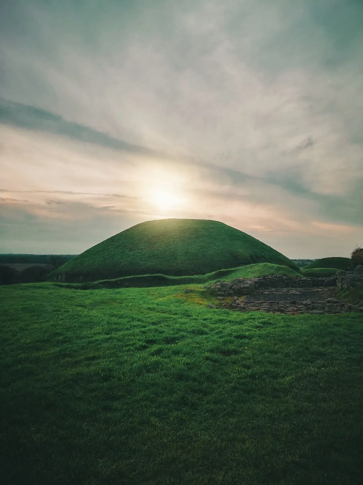 Colina verde sotto un cielo con nuvole e il sole che tramonta dietro di essa, con rovine di pietra in primo piano.