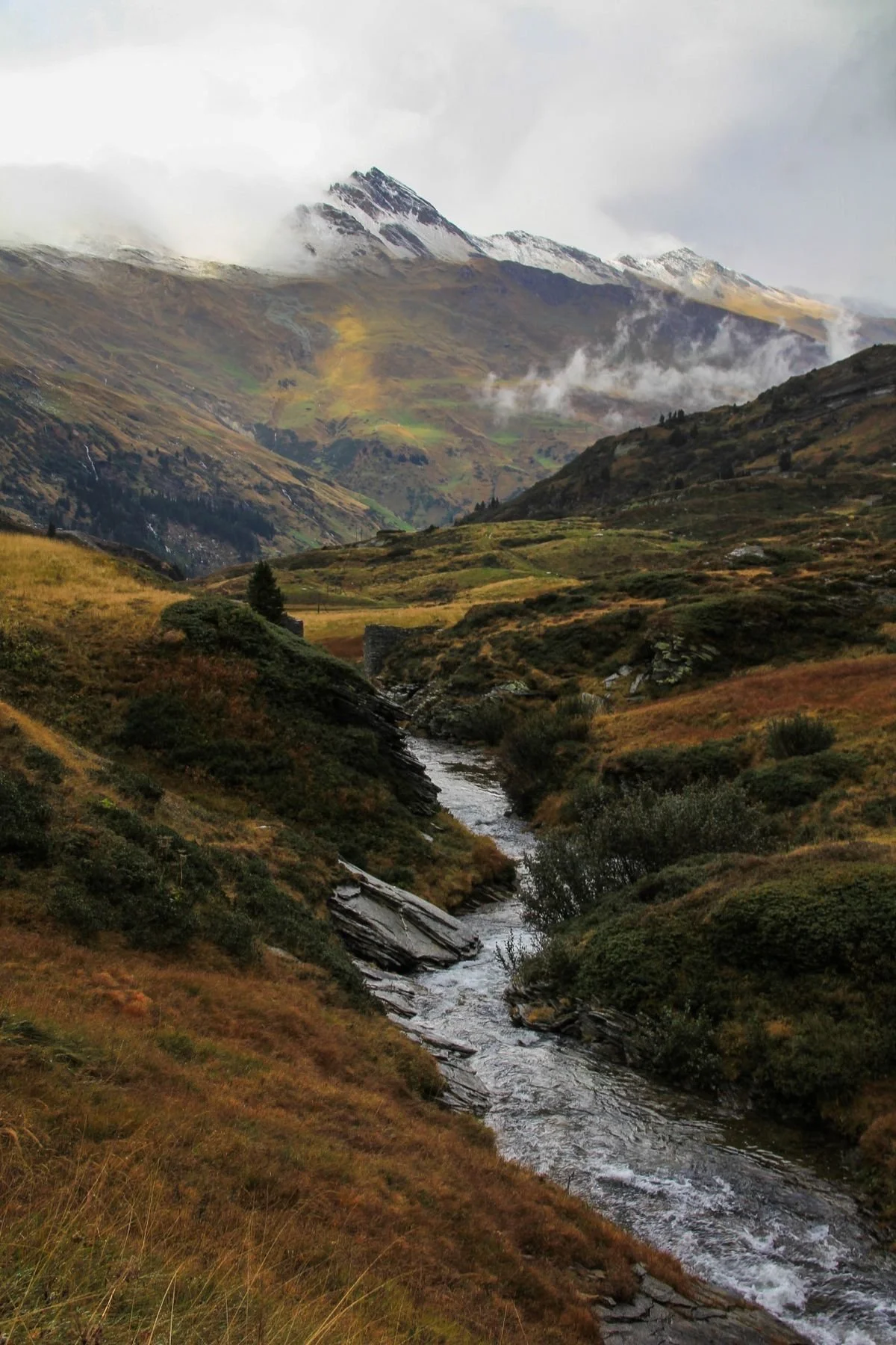 Paesaggio montano con un ruscello che scorre tra colline verdi e montagne con cime innevate e nuvole basse