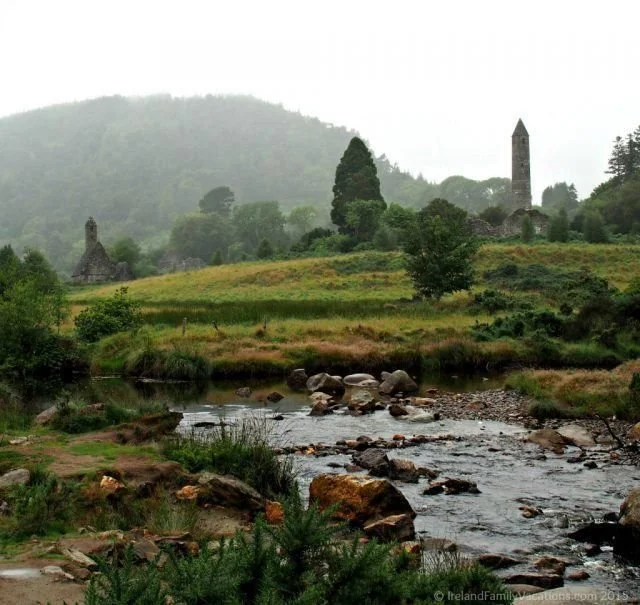 Paesaggio rurale con un fiume, colline verdi, alberi e rovine di un castello con torretta in lontananza, sotto un cielo nuvoloso.