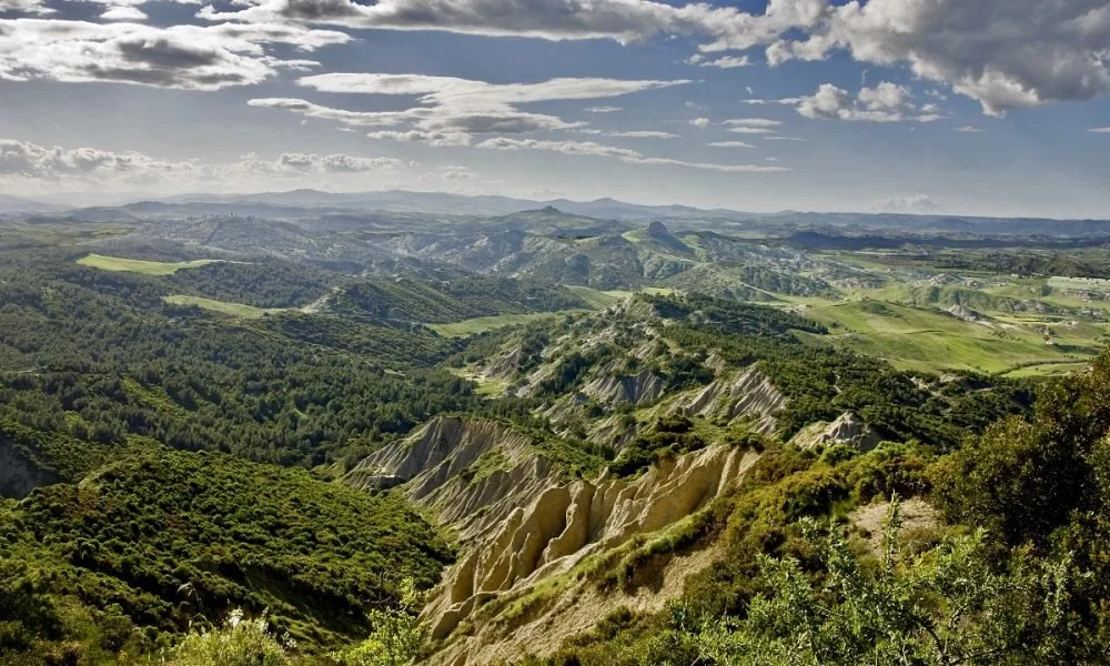 Paesaggio rurale con colline ondulate, vegetazione e nuvole nel cielo.
