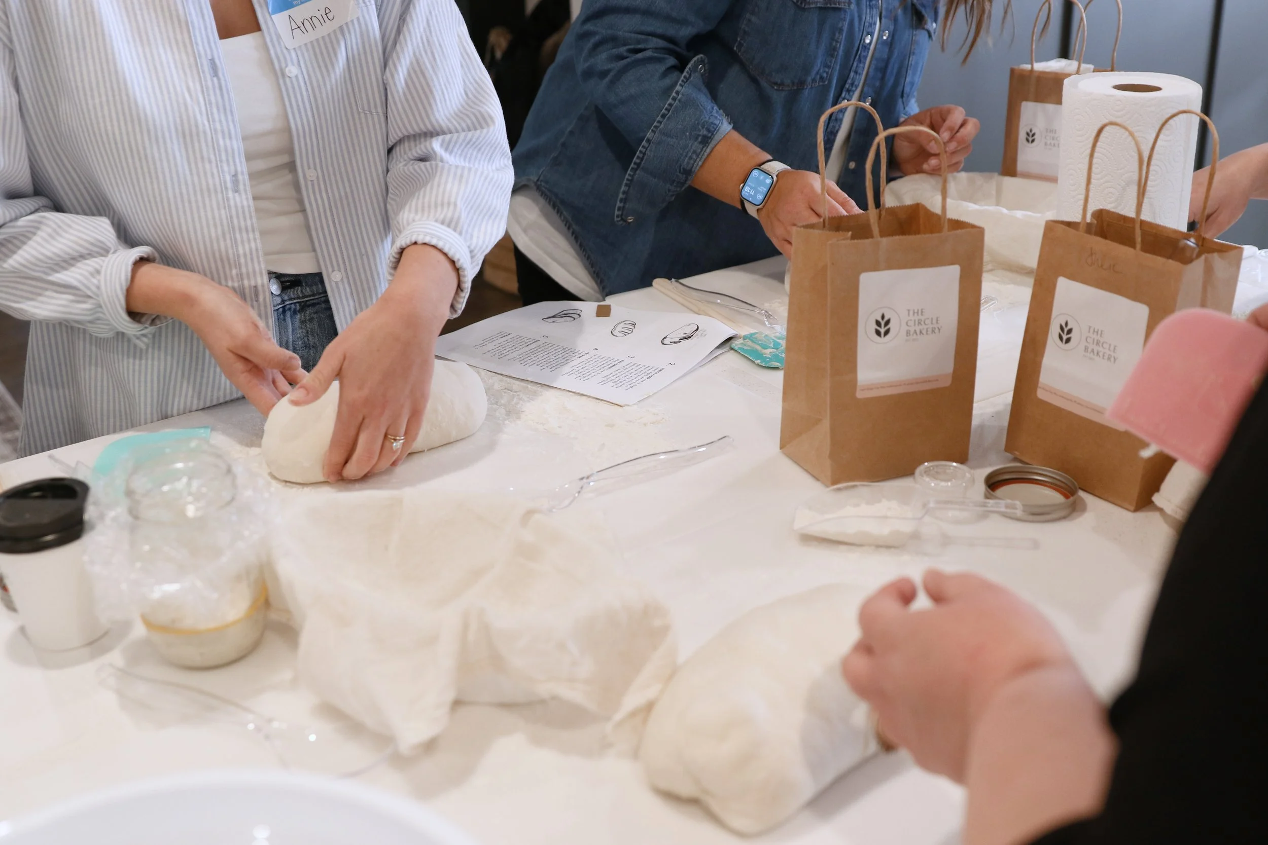 People making bread dough with ingredients and instructions on a table, with paper bags labeled "The Circle Bakery" in the background.
