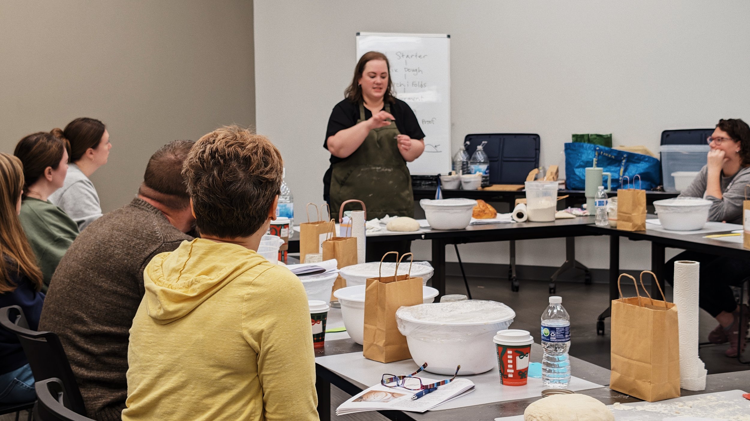 A woman leading a cooking class to a group of students in a classroom with folding tables, pizza dough, ingredients, and paper bags on the tables.