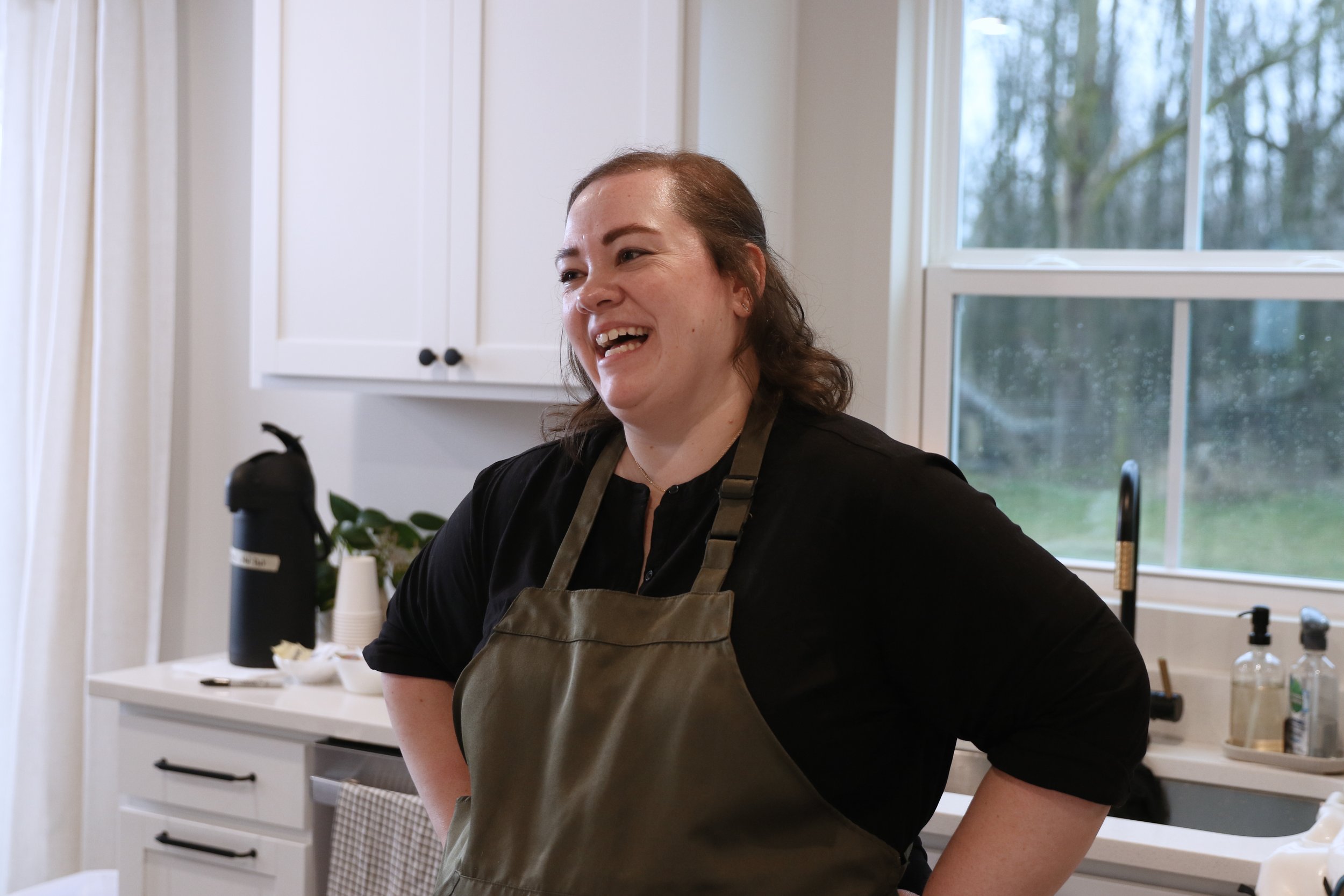A woman smiling in a kitchen wearing a black shirt and an apron, with a window showing trees outside.