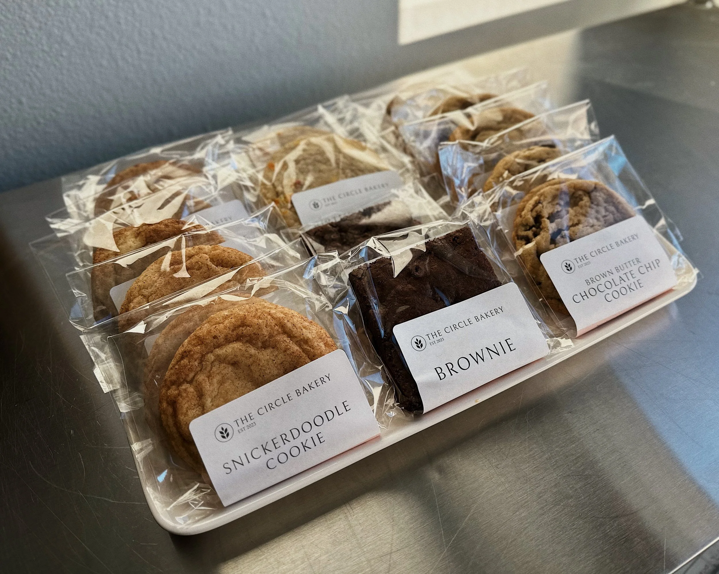 Assorted cookies packaged in clear plastic, labeled with bakery names including 'The Circle Bakery' and cookie types like brownie and snickerdoodle, on a metallic surface.