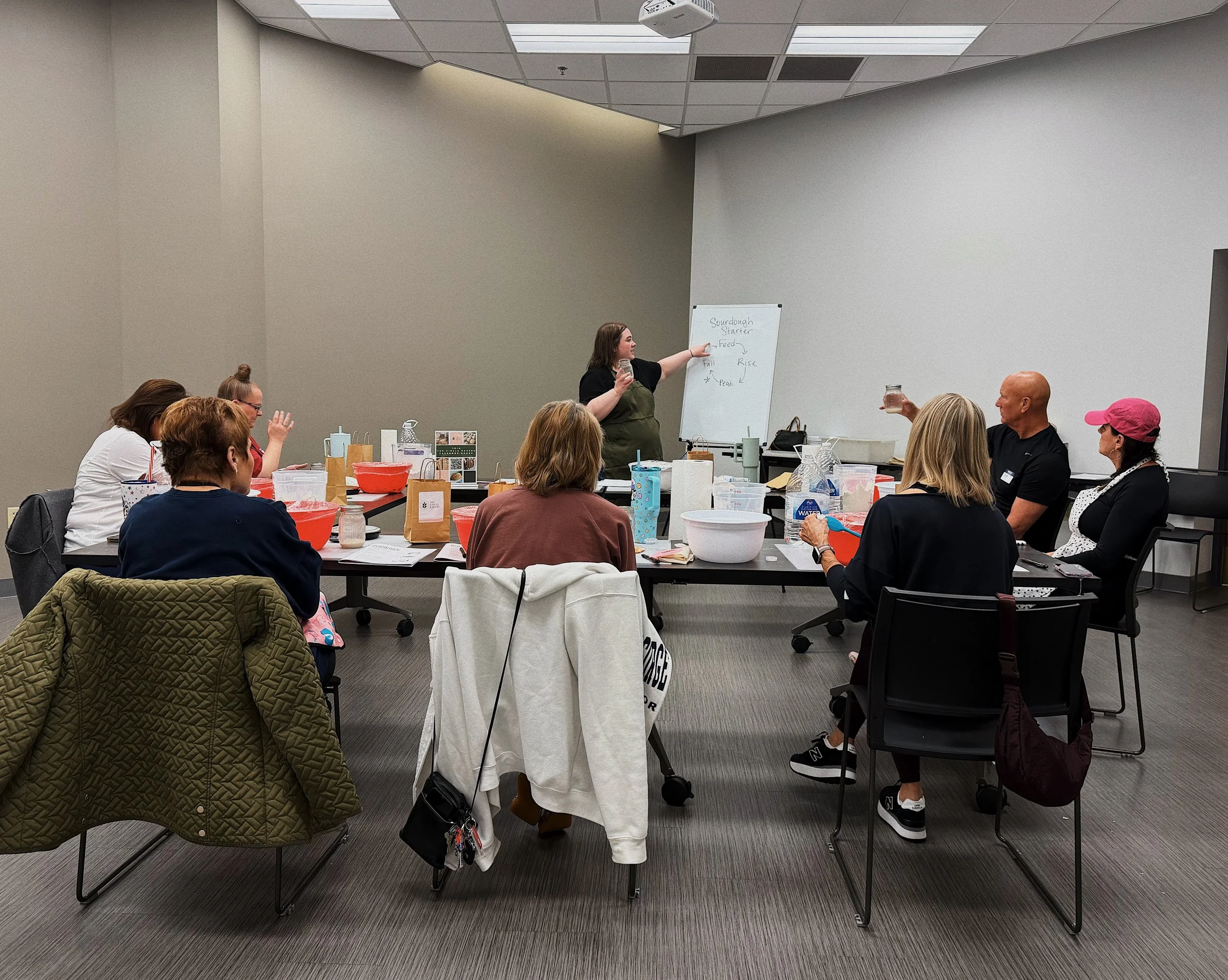 A woman is giving a presentation to a group of people seated at a large table, using a whiteboard with notes about sourdough starter, feed, and peak. The group appears to be engaged, with some holding drinks and taking notes in a conference room.