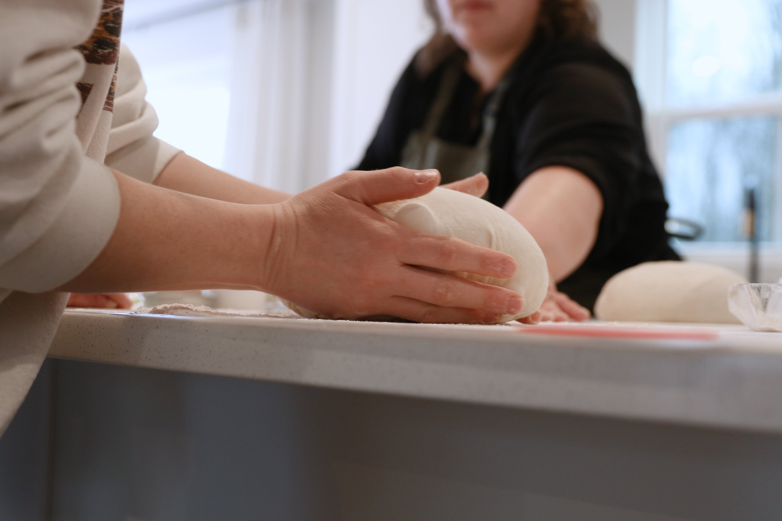 Close-up of hands kneading dough on a white countertop, with a person in a black shirt in the background.