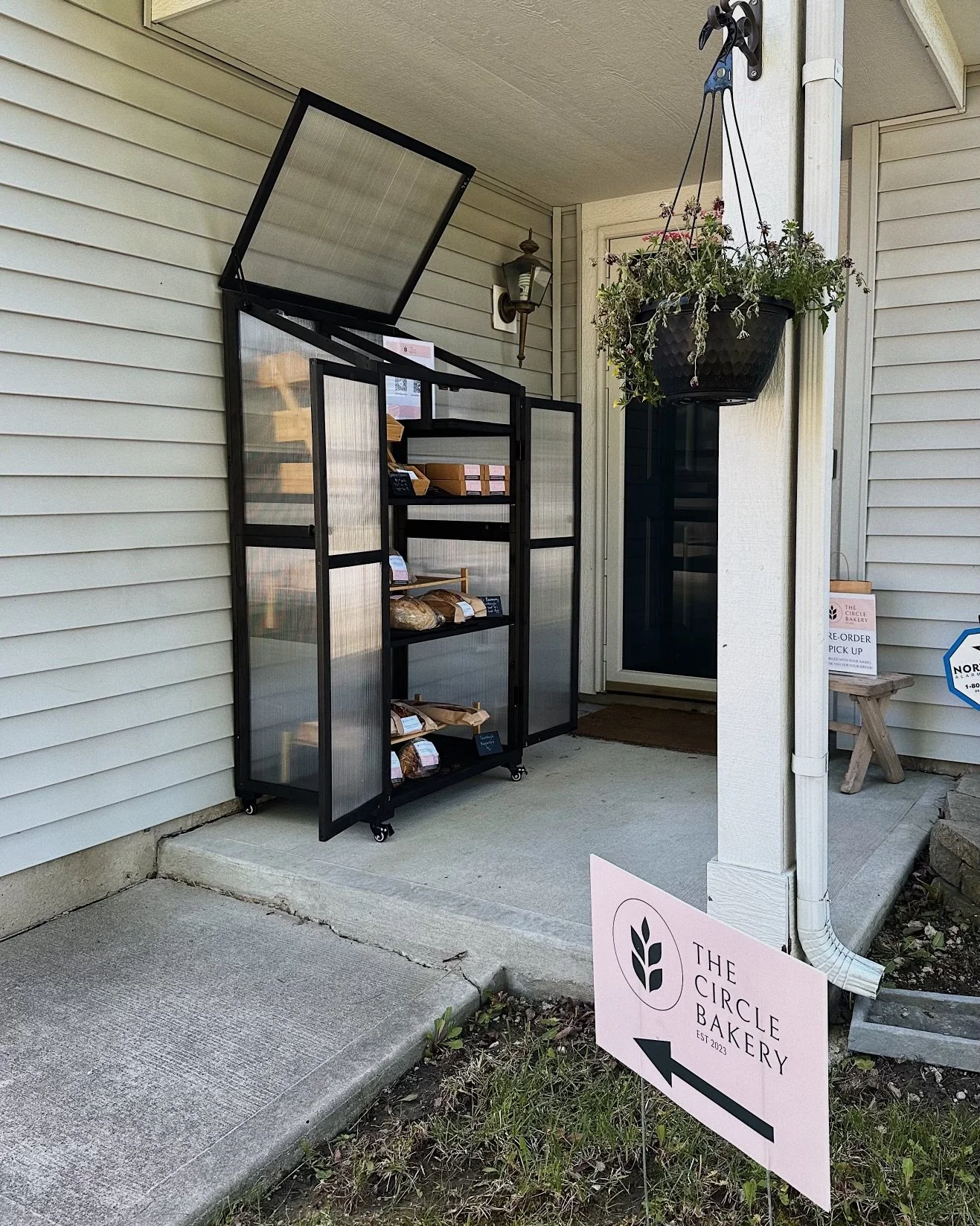 Outside view of a bakery with a sign indicating the bakery is to the left. There is a black metal display case with a glass door and a hinged glass lid, holding baked goods, on a concrete porch next to a white house wall. A potted plant hangs from a bracket above the display case, and a sign on a small stool near the door indicates pre-order pick-up.