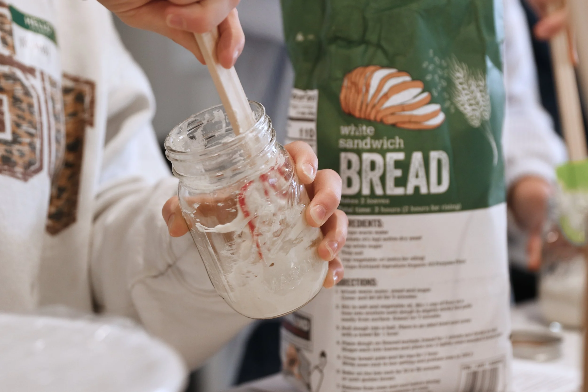 Person pouring sourdough starter from a glass jar, with a green bag of flour in the background.