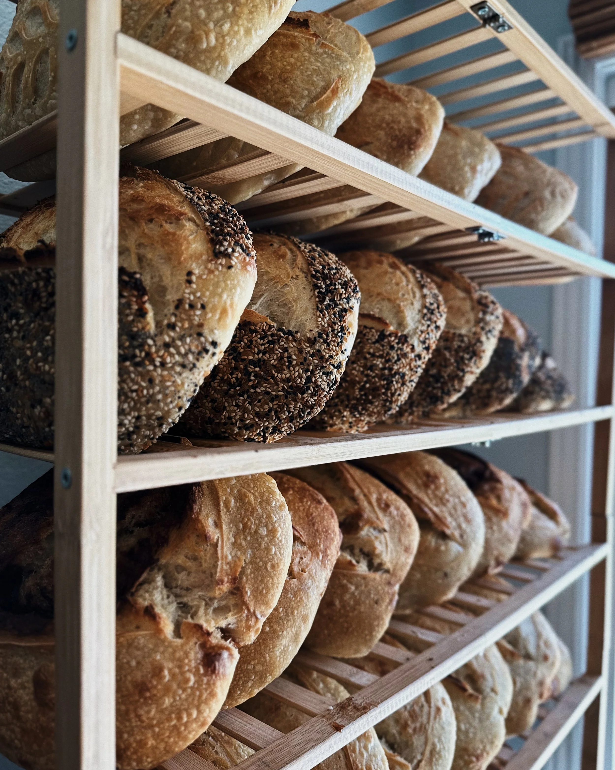 Multiple loaves of freshly baked bread with seeded crusts arranged on wooden racks in a bakery.