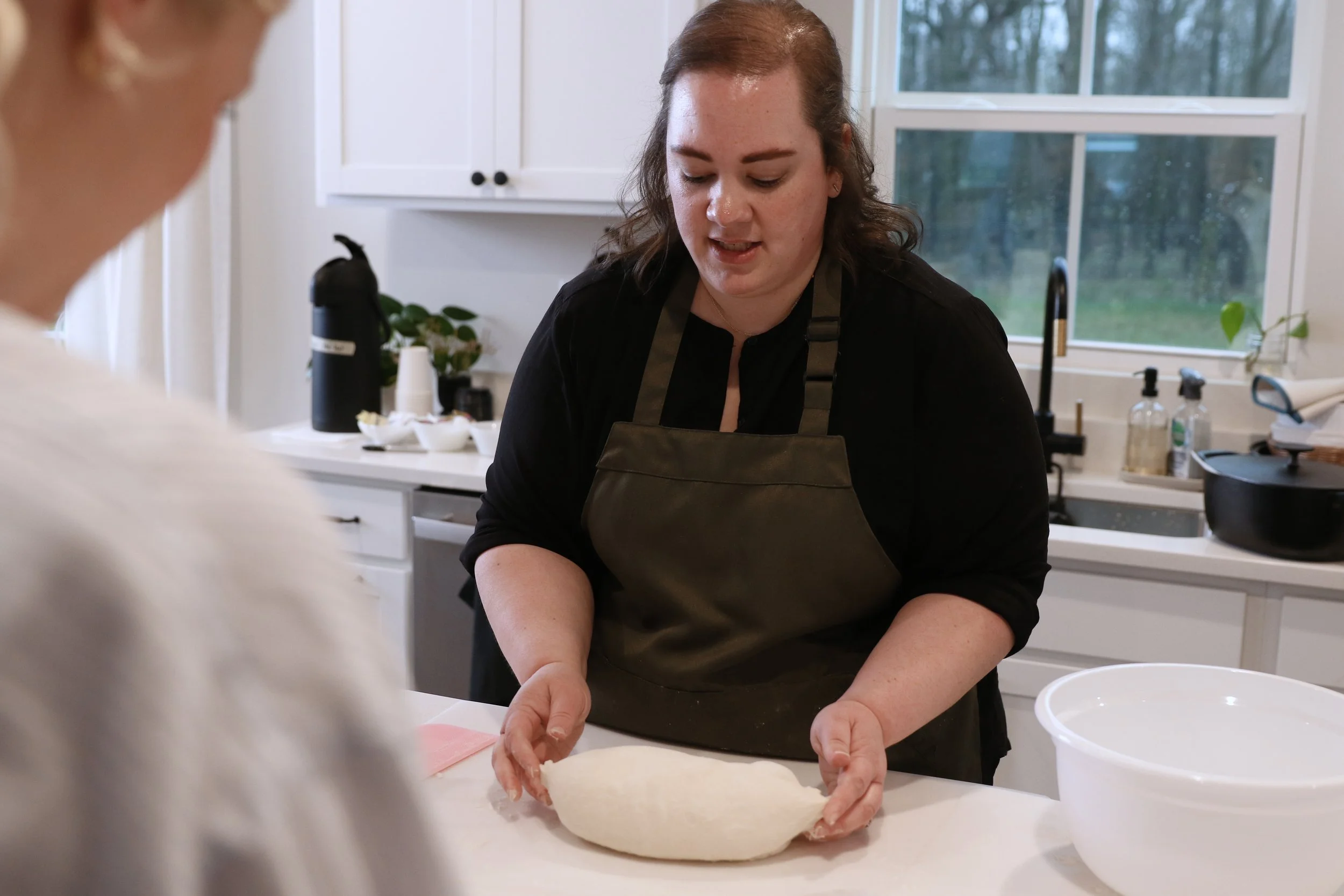 Woman wearing a black apron kneading dough on a white kitchen countertop with a child partially visible in the foreground.