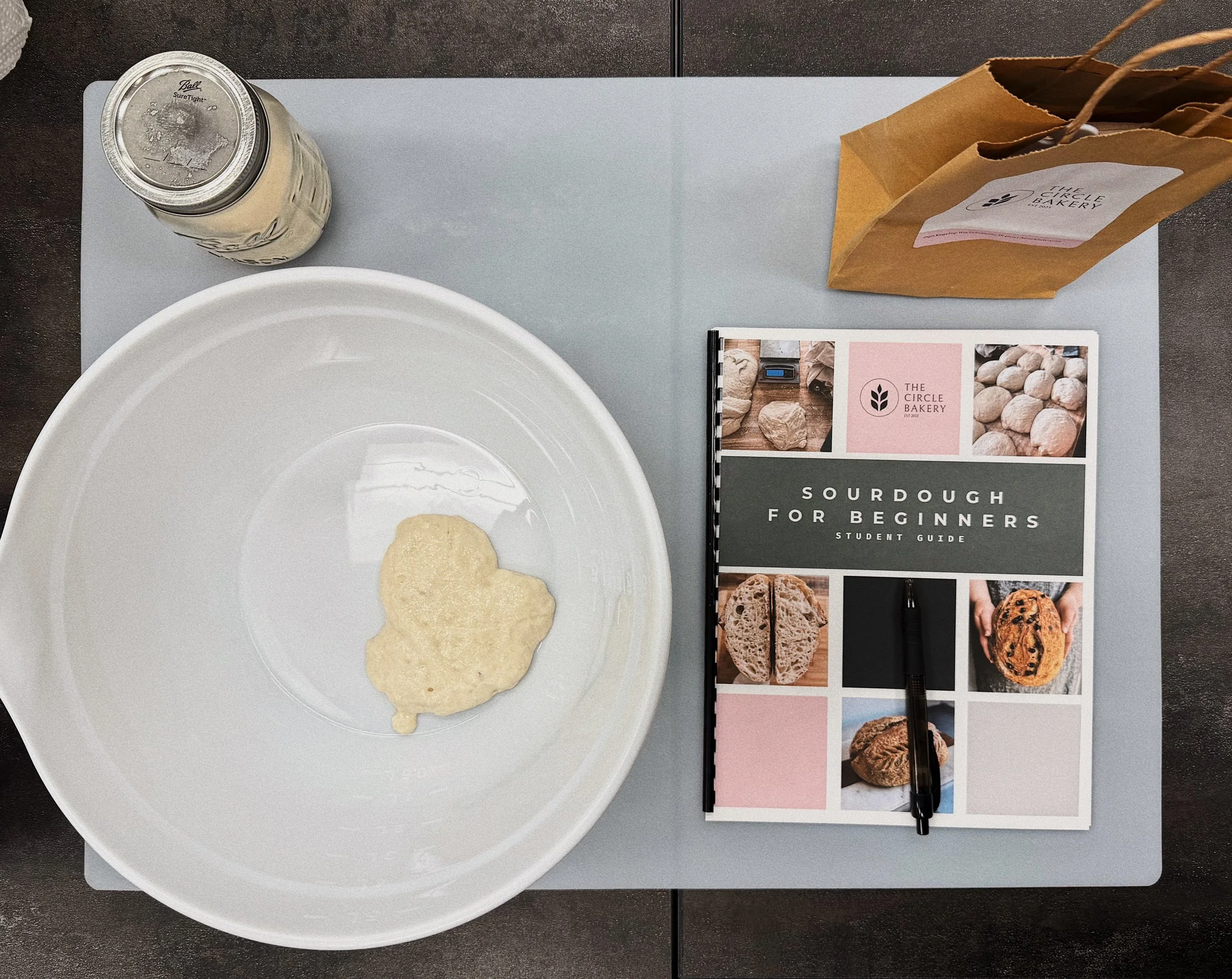 Top view of a gray table with a bowl of sourdough starter, a glass jar of sourdough starter with a metal lid, a brown paper bag from The Circle Bakery, a brochure titled 'Sourdough for Beginners Student Guide,' and a black pen.