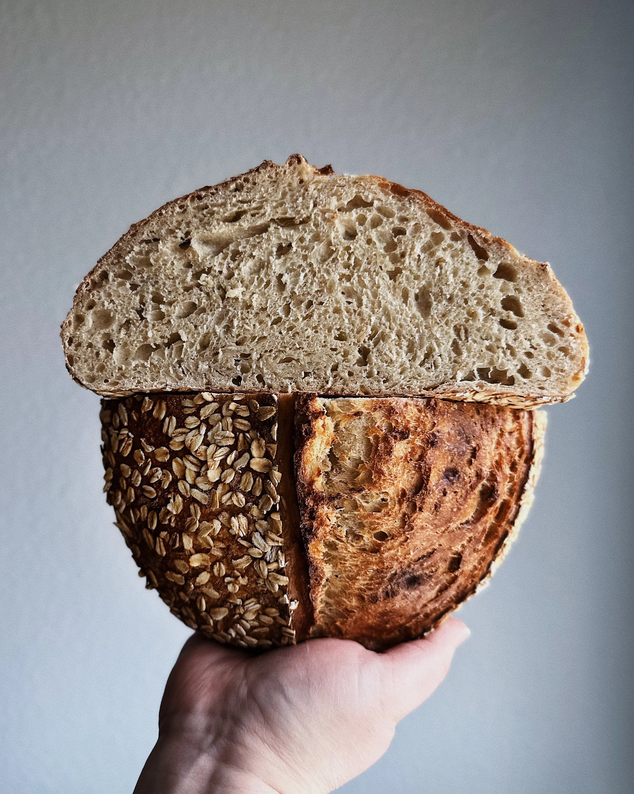 A hand holding a round loaf of bread with oatmeal on the crust, sliced to show the inside texture.