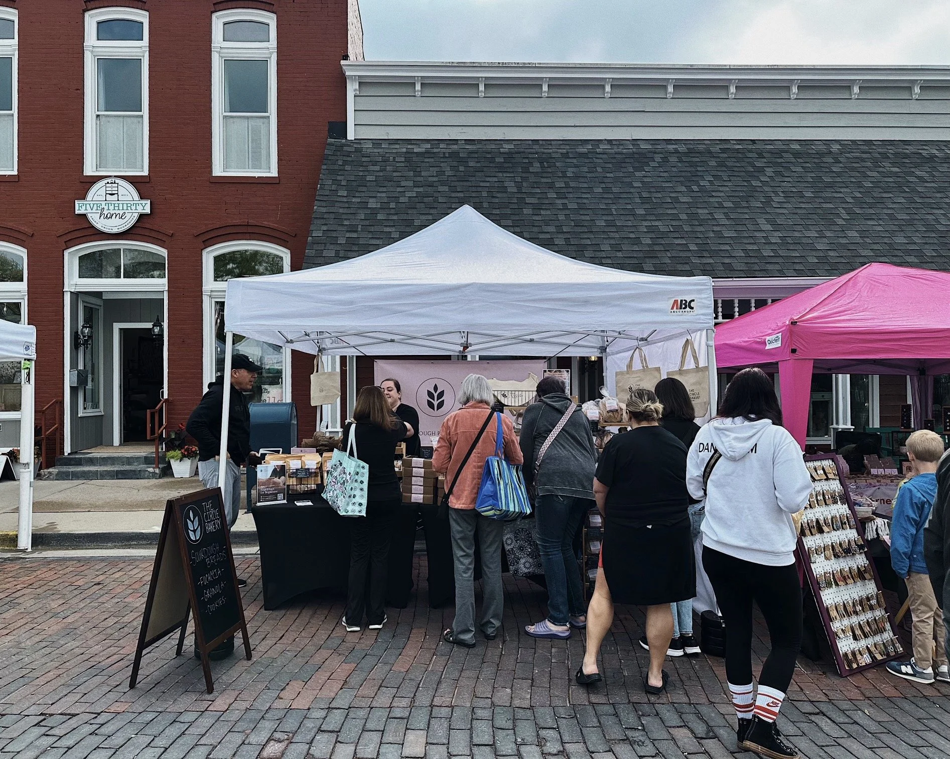 People attending an outdoor market with stalls under white and pink tents in front of a brick building with large windows and a sign that reads "Five Thirty Home."