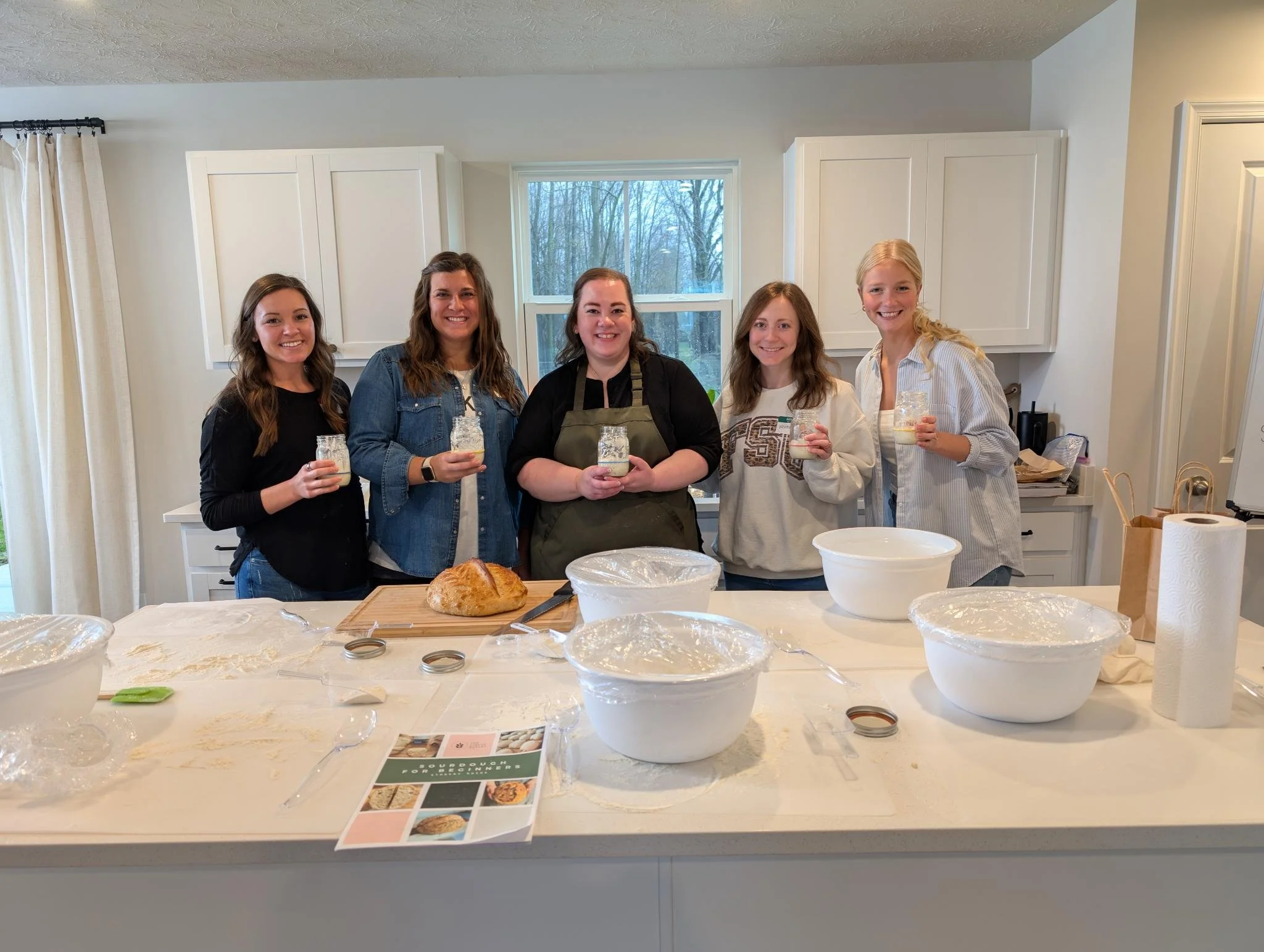 Group of five women after a sourdough workshop, smiling and holding mason jars of sourdough starter, standing in a kitchen behind a table with baking supplies, a loaf of bread, mixing bowls, and utensils.