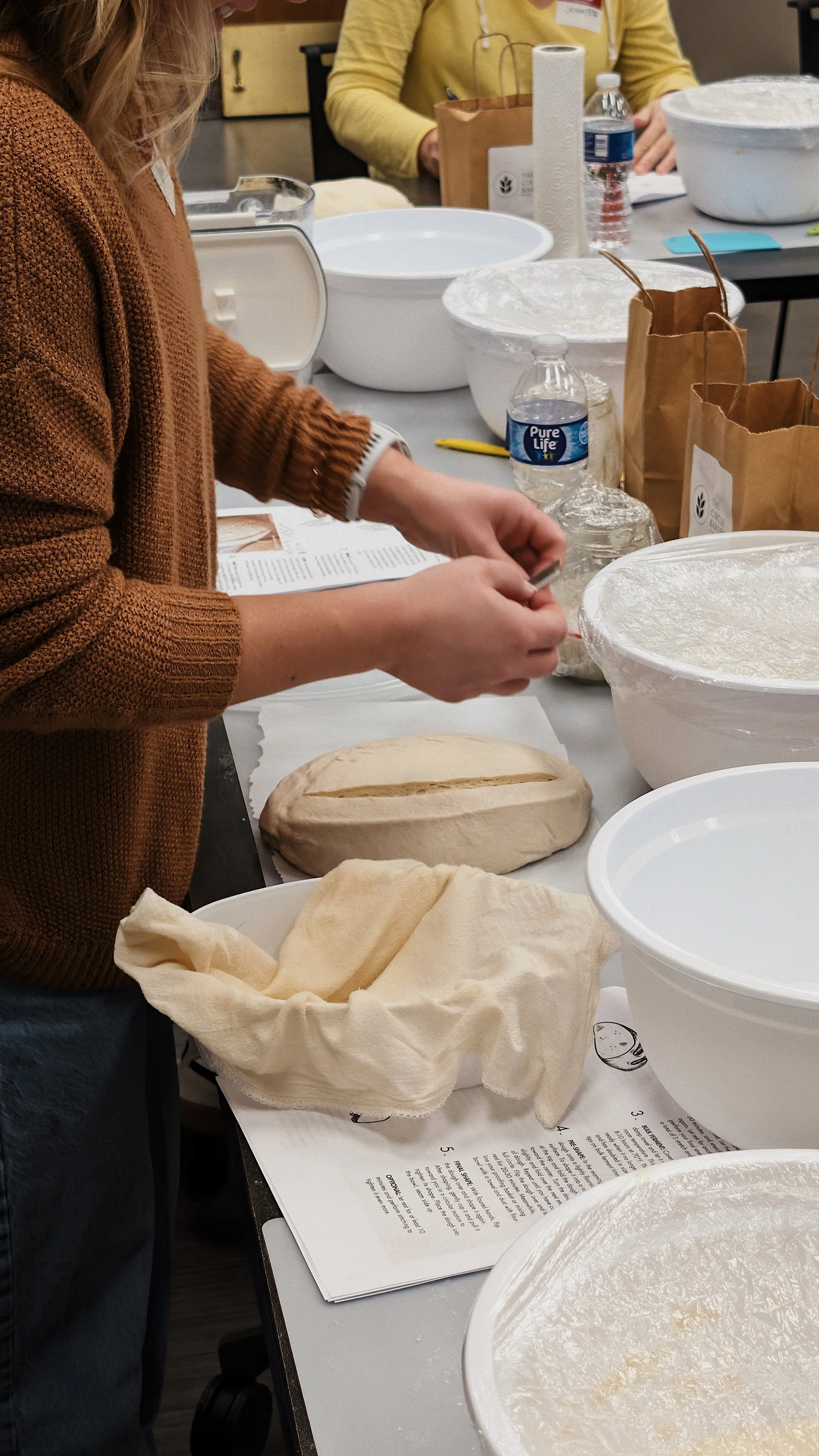 A person with a brown sweater preparing bread dough in a sourdough workshop setting.