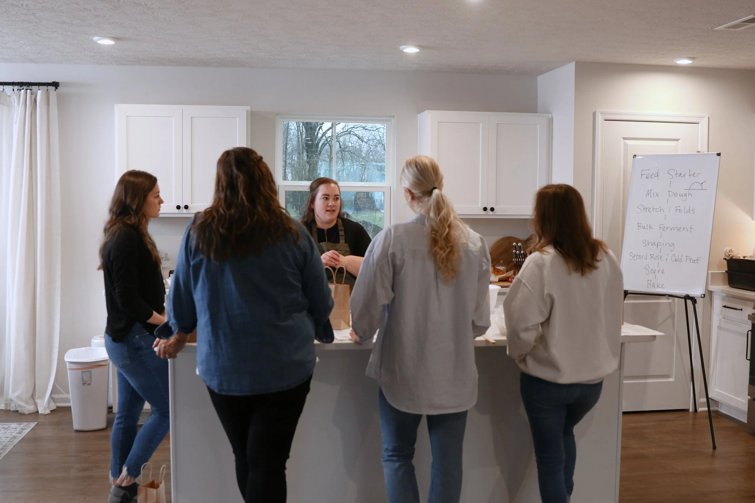 A group of five women gathered around a kitchen island, engaged in conversation during a cooking class or workshop. They are inside a bright, modern kitchen with white cabinets and a window showing trees outside. There is a whiteboard with handwritte