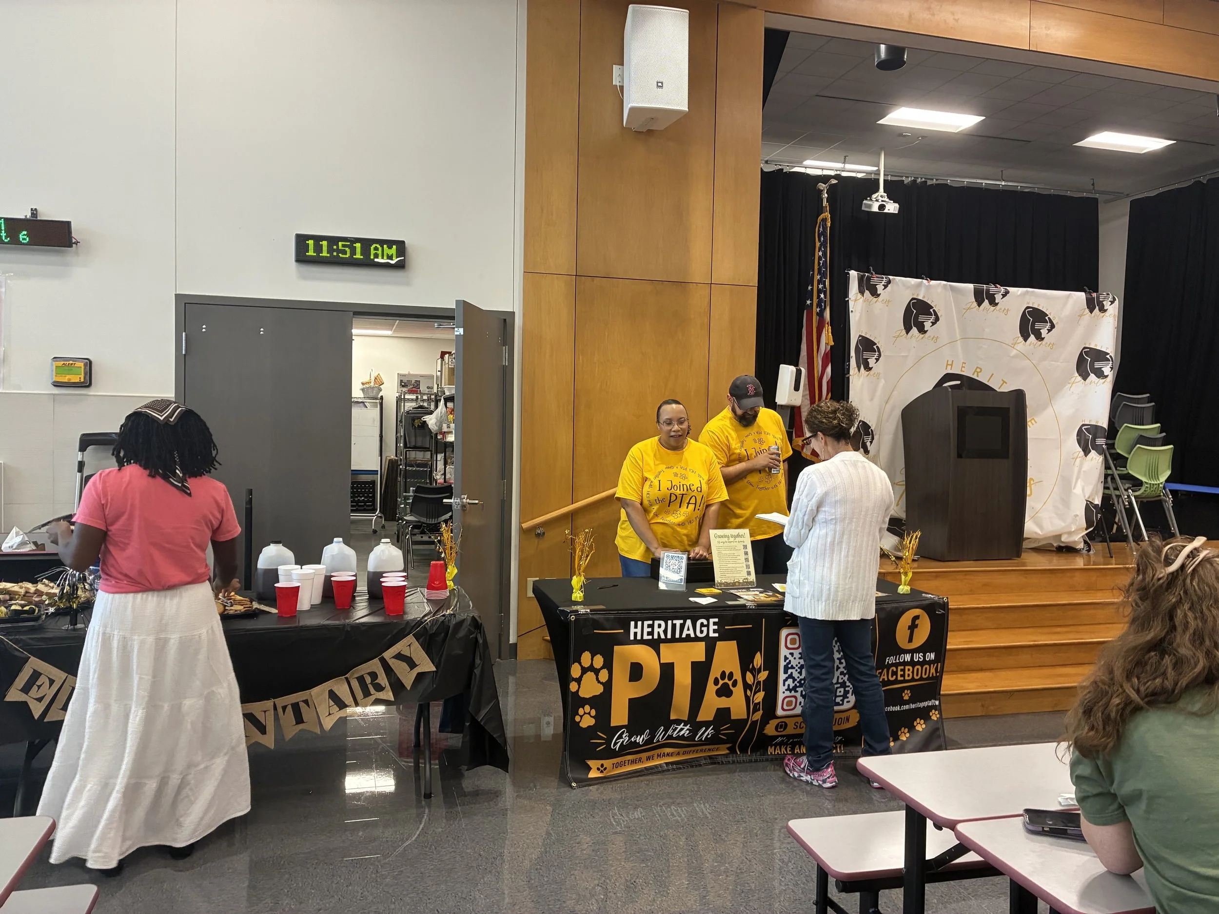 People at a community event, with a table of refreshments and a banner for Heritage PTA. Two women in yellow shirts are at a table and engaged in conversation with a woman in a white sweater. The event appears to focus on community and school involvement.