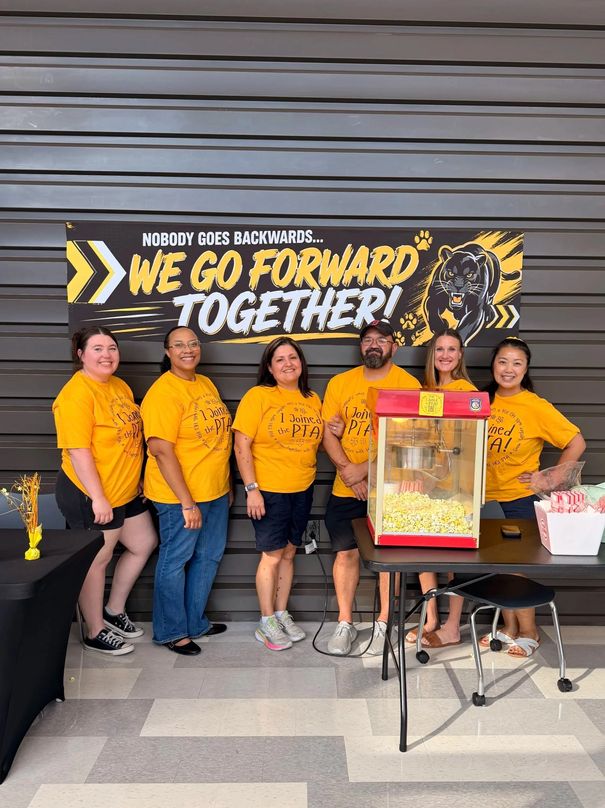 Group of six diverse people wearing yellow shirts standing in front of a black and yellow banner that says 'Nobody goes backwards... We go forward together!' with a panther graphic. They are smiling and posing for a photo at a school or community event. There is a popcorn machine on a table in front of them.