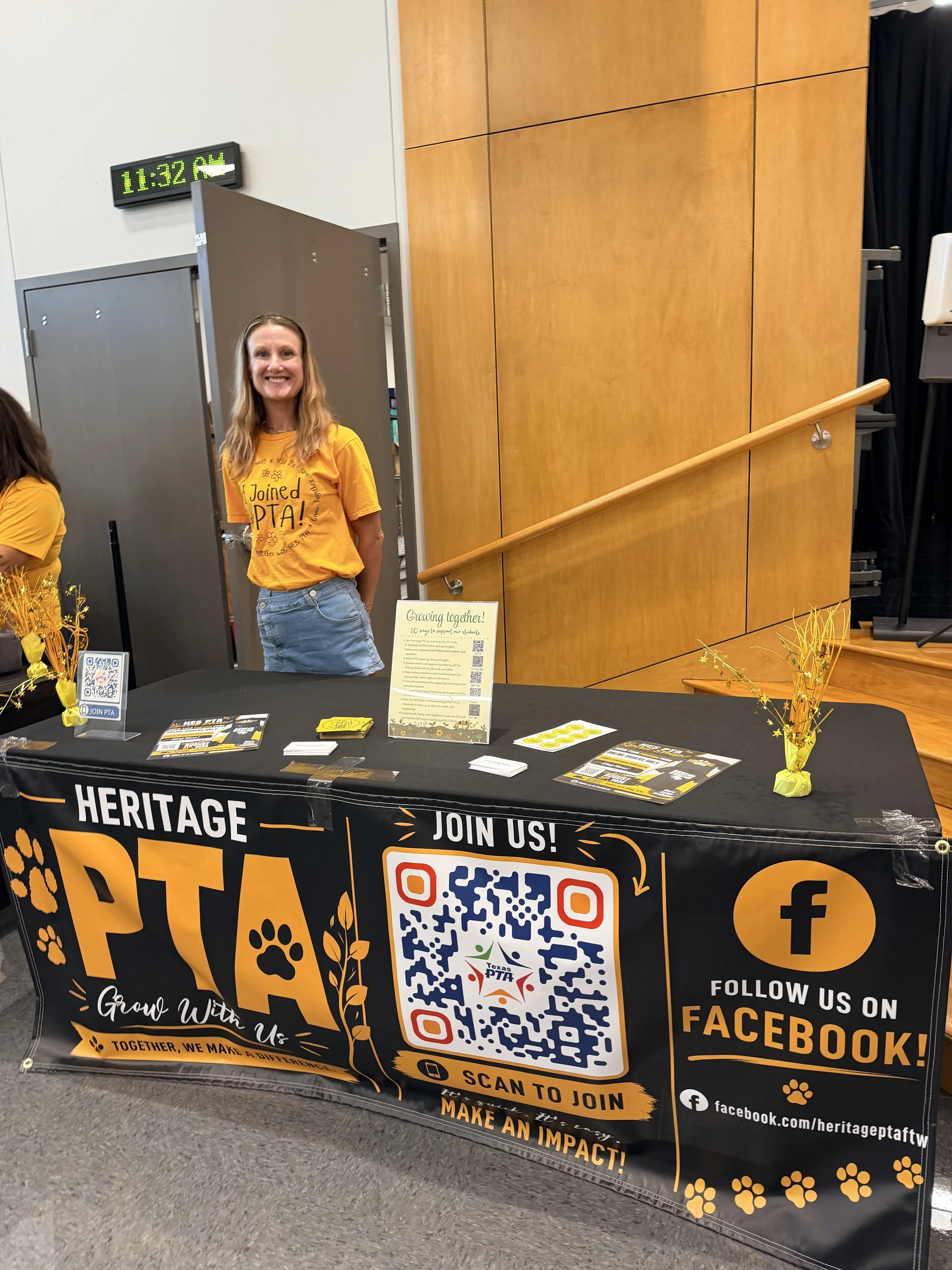 A woman standing behind a table at a Heritage PTA event, smiling. The table has a black and orange tablecloth with a QR code, social media info, and promotional text. Behind her is a wooden wall, and there is a sign that says "Giving together!" on the table.