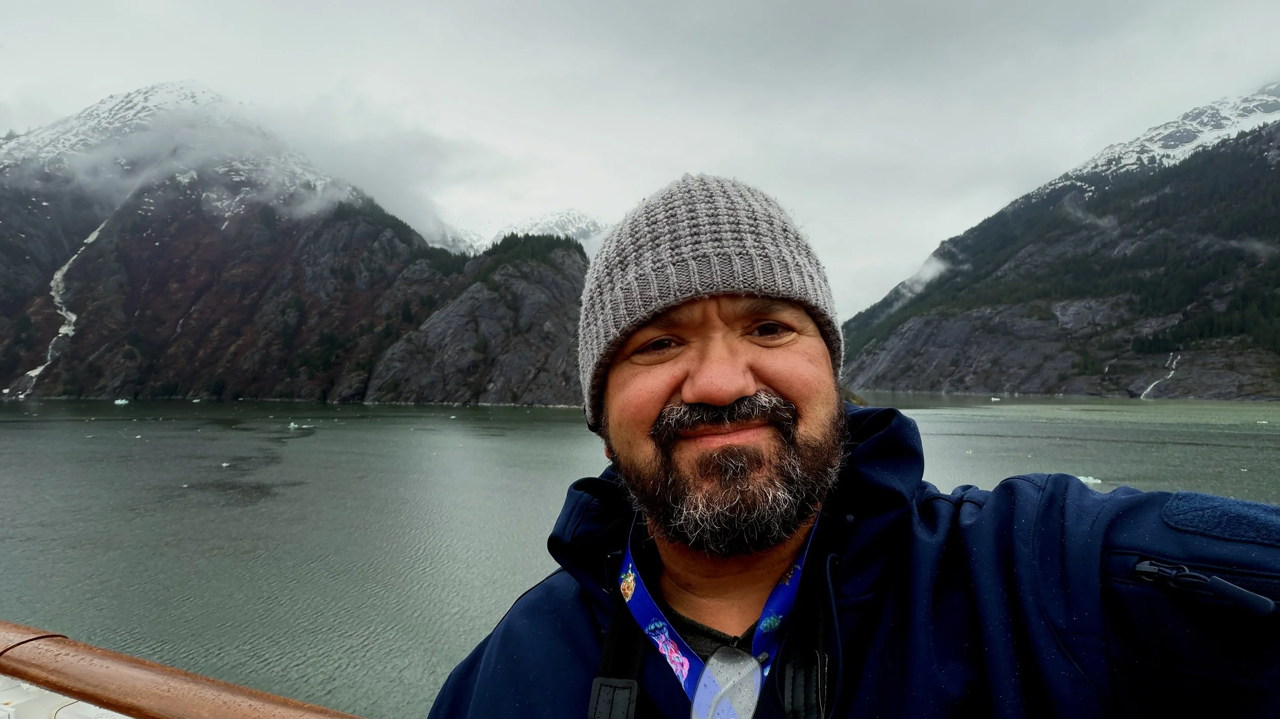 A man with a gray knit beanie and a beard taking a selfie with a mountain lake and snow-capped mountains in the background.