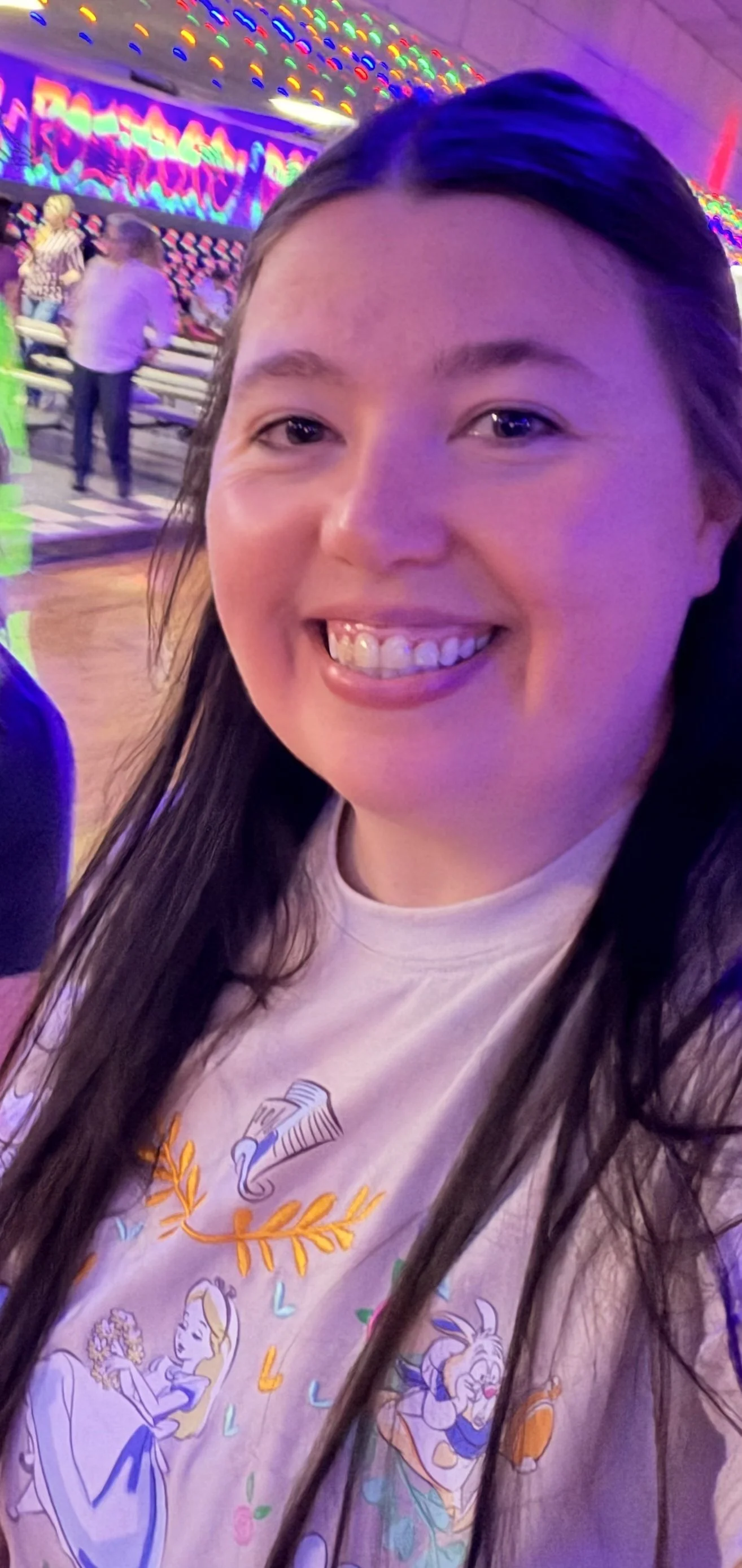 A young woman smiling at an arcade or entertainment venue with colorful neon lights and people in the background.