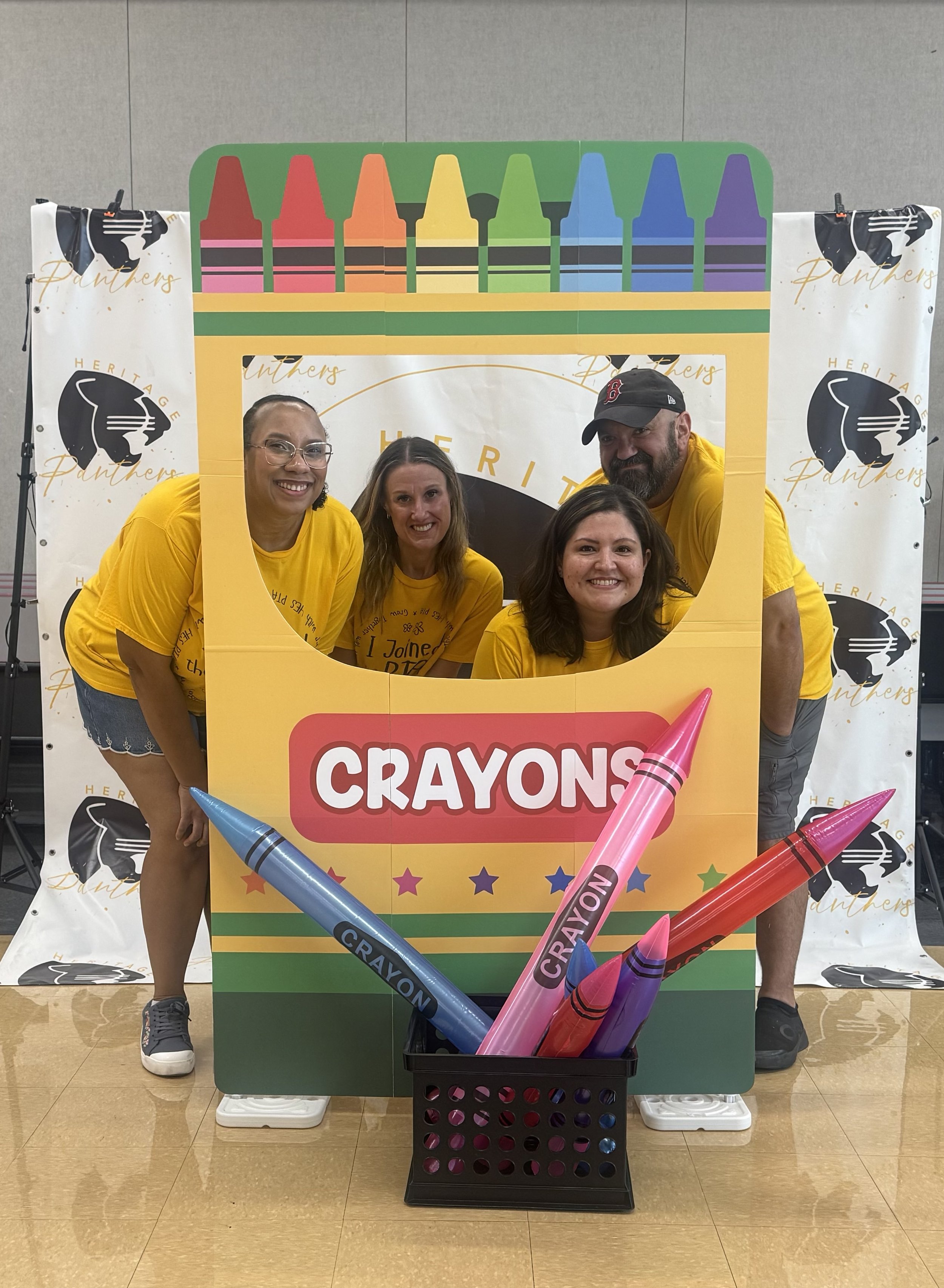 Four people wearing yellow shirts posing behind a large cutout of a crayon box at a event, with oversized crayons in front, and a colorful backdrop with crayons at the top.
