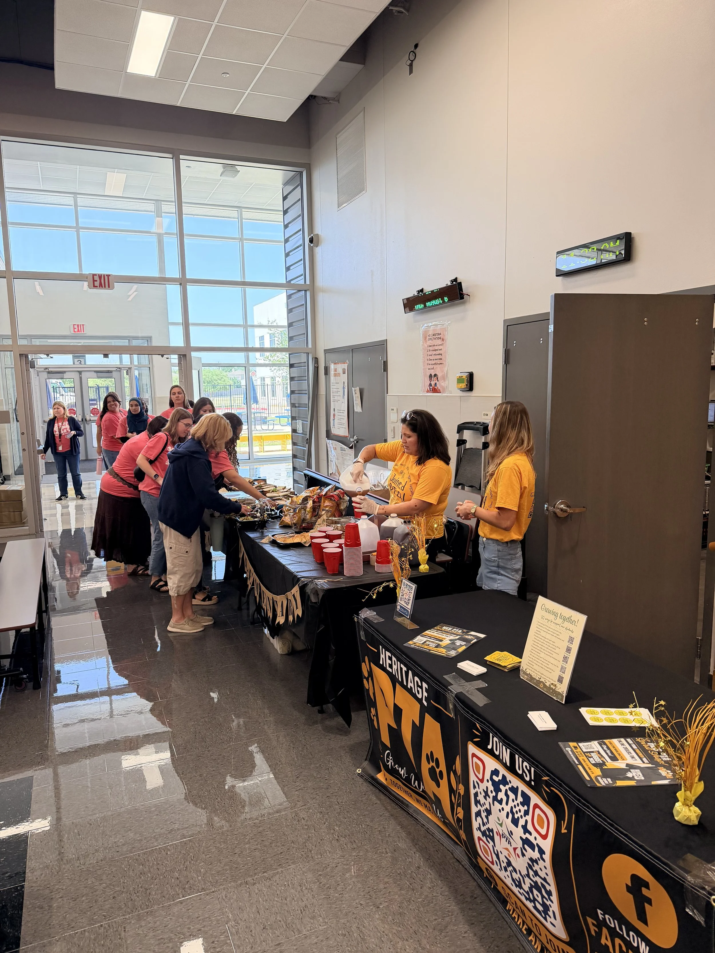 People lining up at a donation event table inside a building with large glass windows showing the outside with blue skies. The table has various flyers, a QR code, and donation information. Several volunteers in yellow and pink shirts are assisting attendees.