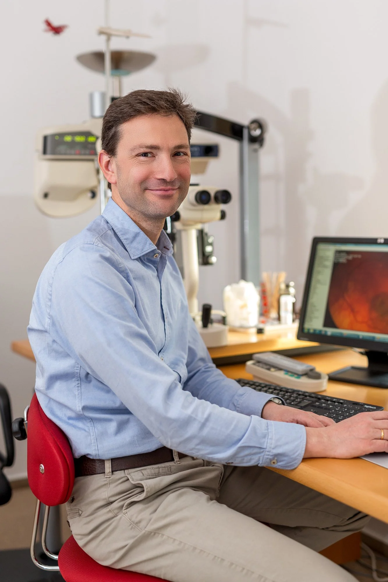 Ein Mann sitzt an einem Schreibtisch mit einem Computer, auf dem ein Bild eines Auges zu sehen ist, in einem medizinischen Untersuchungsraum mit medizinischer Ausrüstung im Hintergrund.