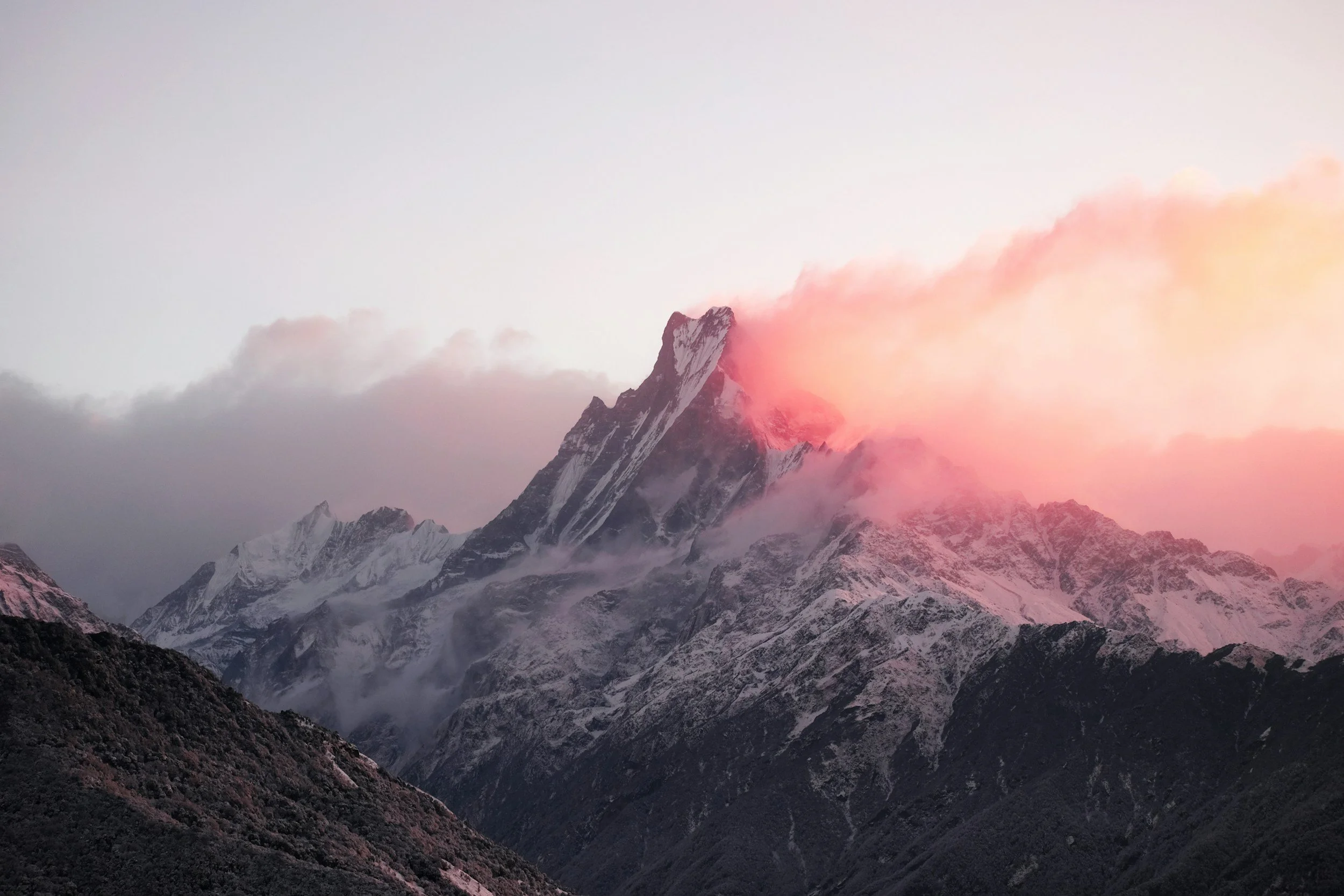 Snow-covered mountain peaks at sunset with pink and orange hues in the sky and billowing clouds around the peaks.
