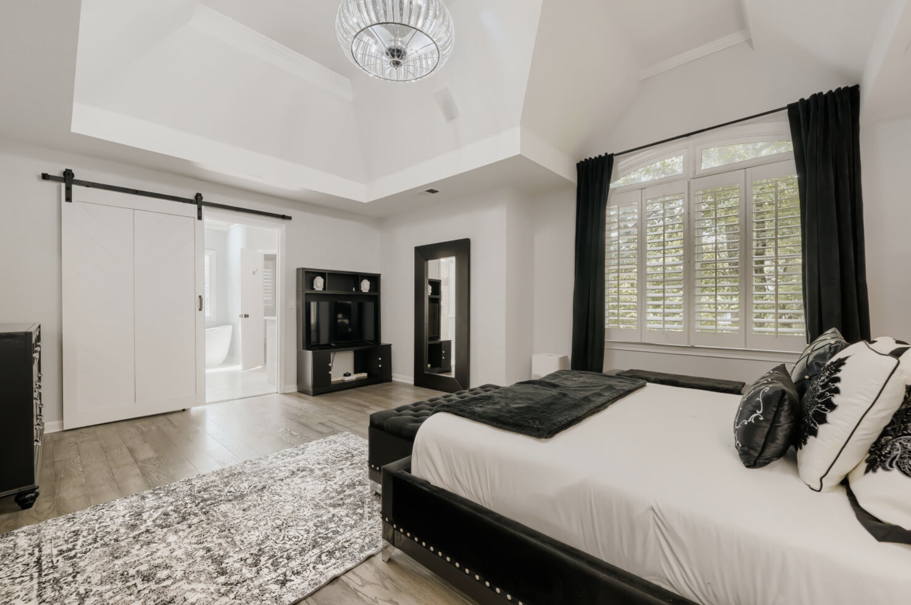 A bedroom interior with a large bed with black and white bedding, black pillows, and a dark headboard. There is a large window with white shutters and black curtains. A black TV stand with a flat-screen television is on the wall, and a tall mirror is next to it. A sliding barn door leads to an adjacent bathroom with a white bathtub. The room has wood flooring and a gray patterned area rug.